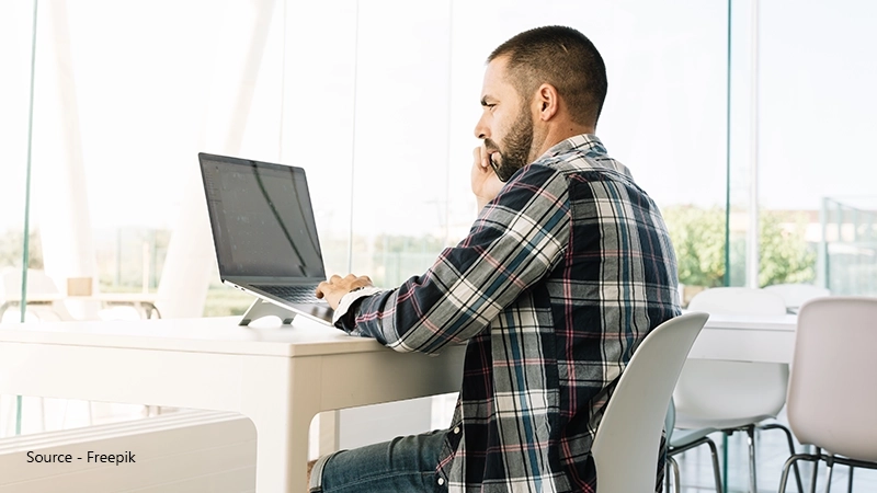 A man with a beard sits at a modern, brightly lit office desk, working intensely on a laptop, emphasising eye strain from prolonged VDU usage.