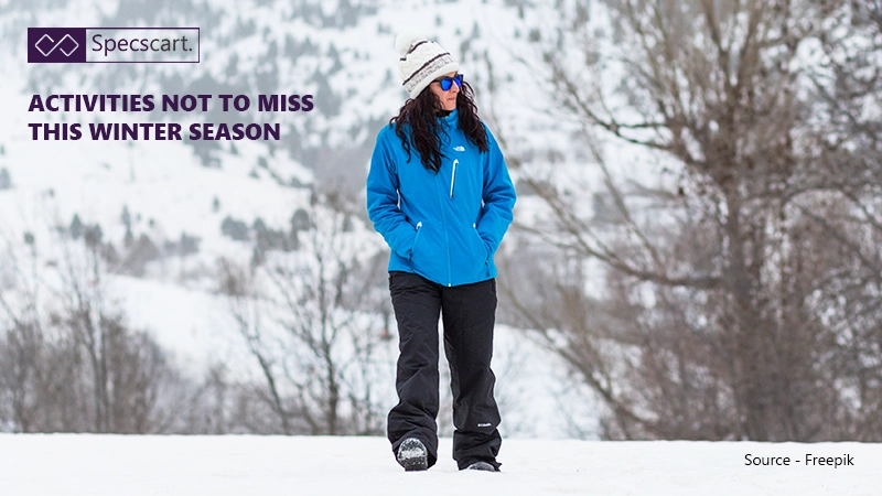 A woman in a bright blue winter jacket, black ski pants, and a hat walks across a snow-covered landscape. She's wearing sunglasses and looks ready for winter activities.