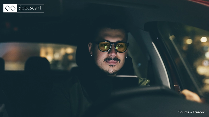 A driver sits in a dark car wearing yellow-tinted glasses, demonstrating how these lenses are used to reduce glare and improve contrast at night.