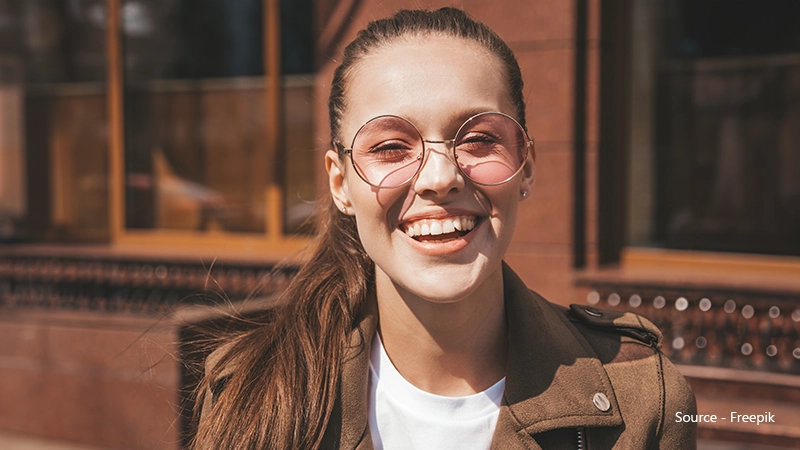 A young woman is outdoors on a sunny day, wearing a brown jacket and round, pink-tinted sunglasses with thin metal frames, smiling broadly with her eyes closed.