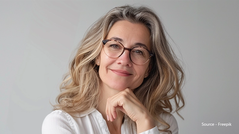 A smiling woman with long, wavy blonde and grey hair poses with her chin resting on her hand. She wears a white collared shirt and round, tortoiseshell browline glasses.