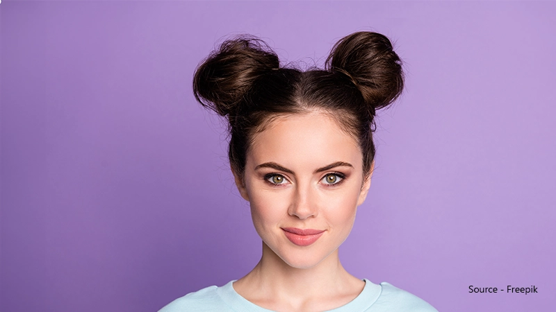 A woman with brown hair styled in two large space buns smiles slightly, wearing a light blue shirt against a lavender background.