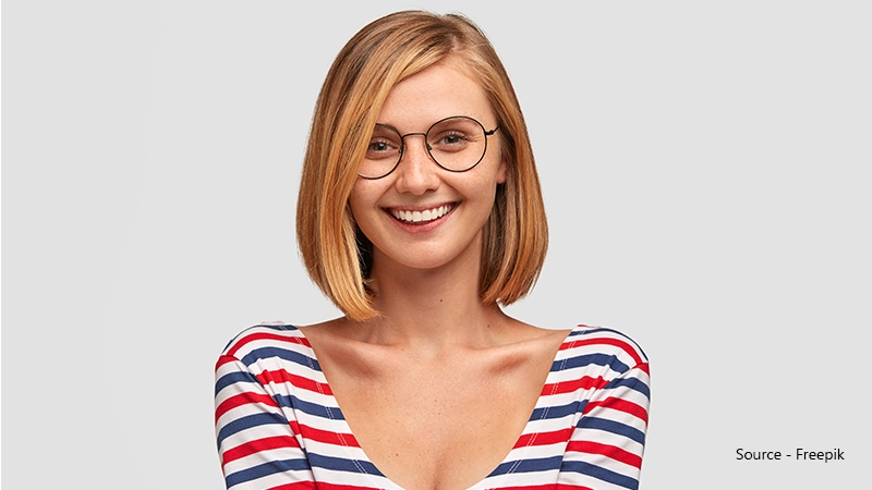 A young woman with short blonde hair and freckles smiles widely at the camera. She wears a striped red, white, and blue top and round, thin black-rimmed glasses.