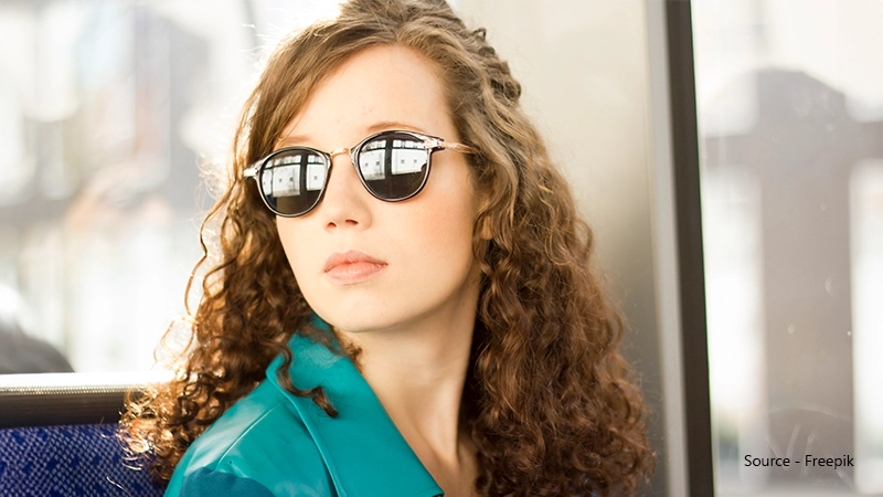 A woman with long, curly brown hair and a turquoise leather jacket looks away from the camera. She wears round sunglasses with dark lenses and gold accents.