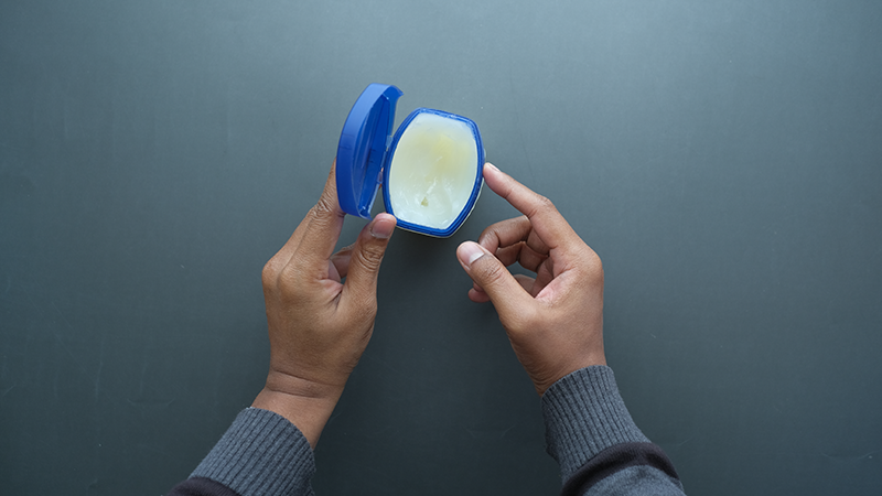 Overhead view of hands opening a jar of petroleum jelly, used as a simple method for removing stubborn eye makeup.