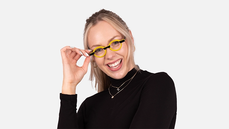 Smiling woman in a black turtleneck wearing bold yellow-rimmed round glasses, radiating cheerfulness and creative energy.