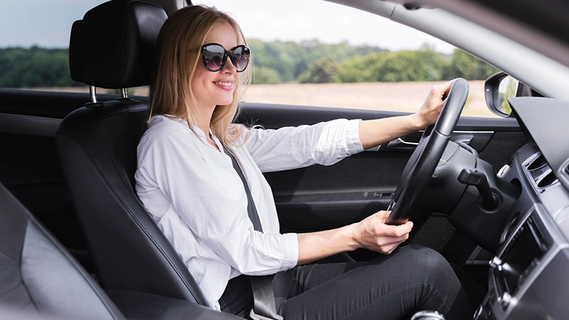 A smiling woman driving a car while wearing oversized gradient sunglasses, highlighting ideal eyewear for driving.