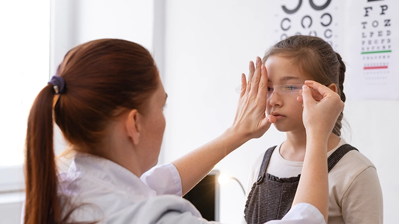 Optometrist measuring a young girl's eyes with a transparent ruler during an exam in a bright clinic room.