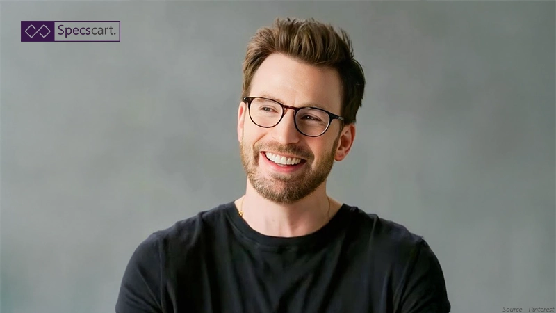 A smiling Chris Evans with styled hair and a beard, wearing classic dark round-frame glasses and a black t-shirt against a grey background.