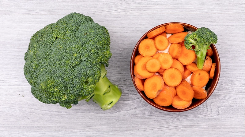 Fresh broccoli next to a bowl of sliced carrots, highlighting vitamin-rich foods that support healthy eyes.