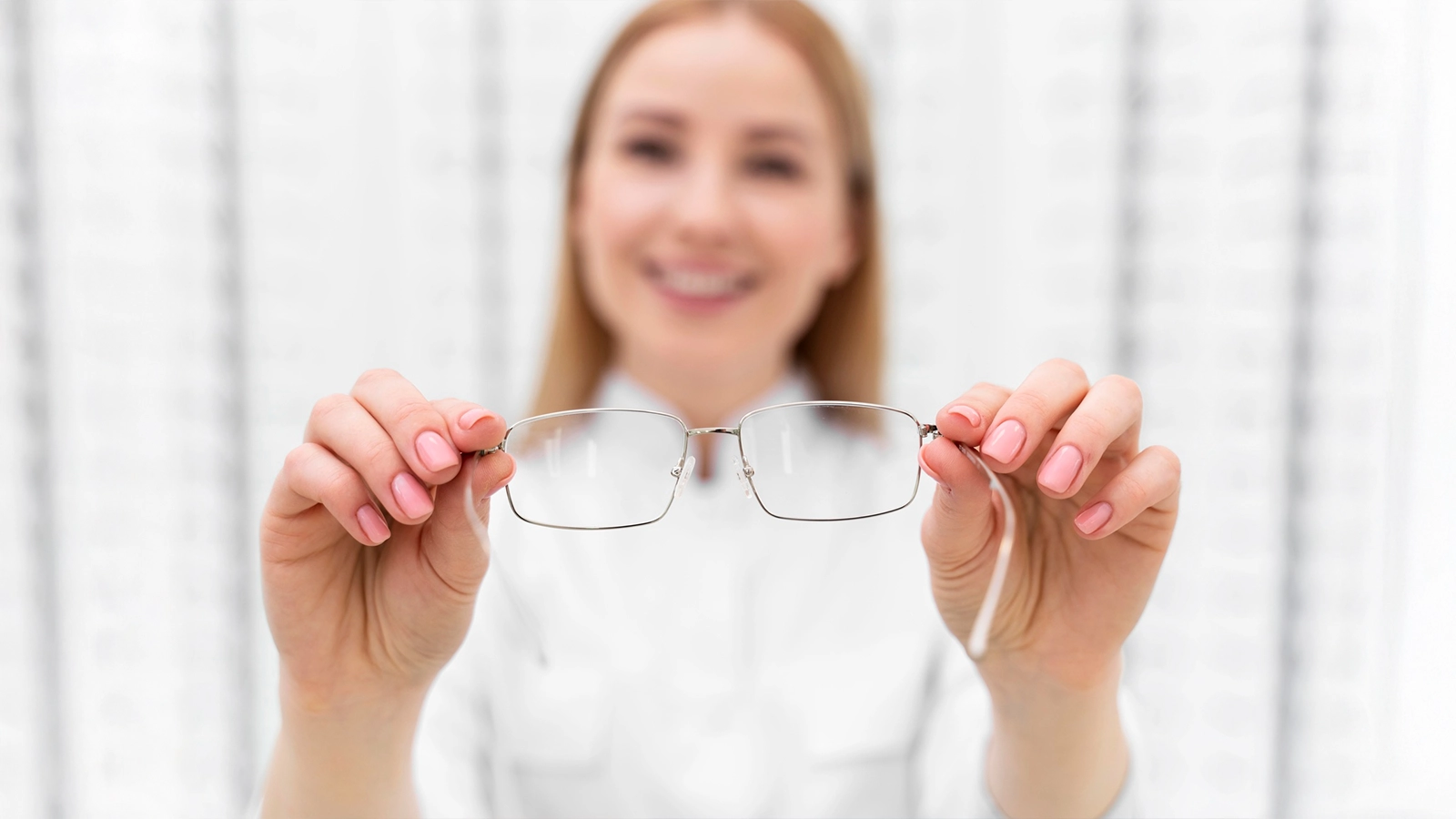A blurred optician in the background holding a pair of thin silver rectangular glasses toward the camera, focusing on the clear lenses and frame.