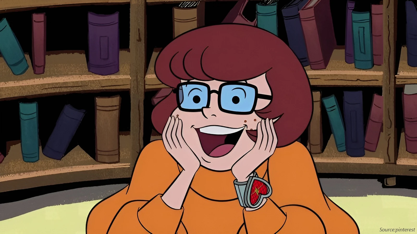 Velma Dinkley, a cartoon character, smiles with her hands on her cheeks. She wears a bright orange turtleneck, square black glasses, and a watch with a red heart-shaped cover against a bookshelf background.