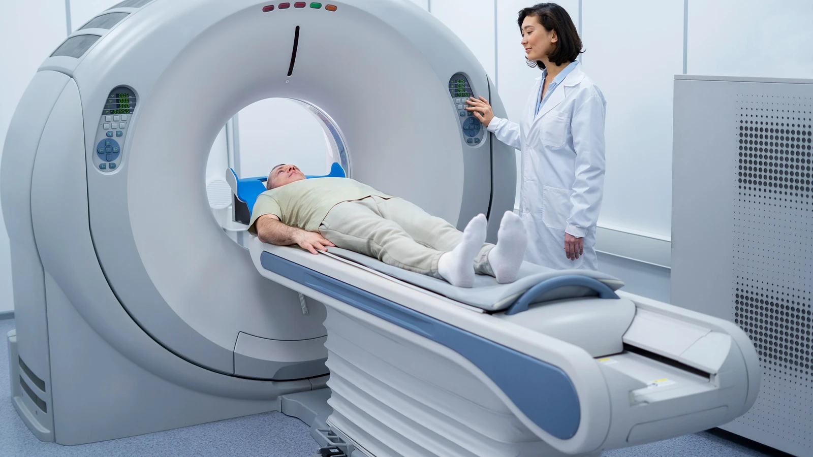 A patient lying on a motorised bed being prepared for an MRI or CT scan by a medical professional in a lab coat.