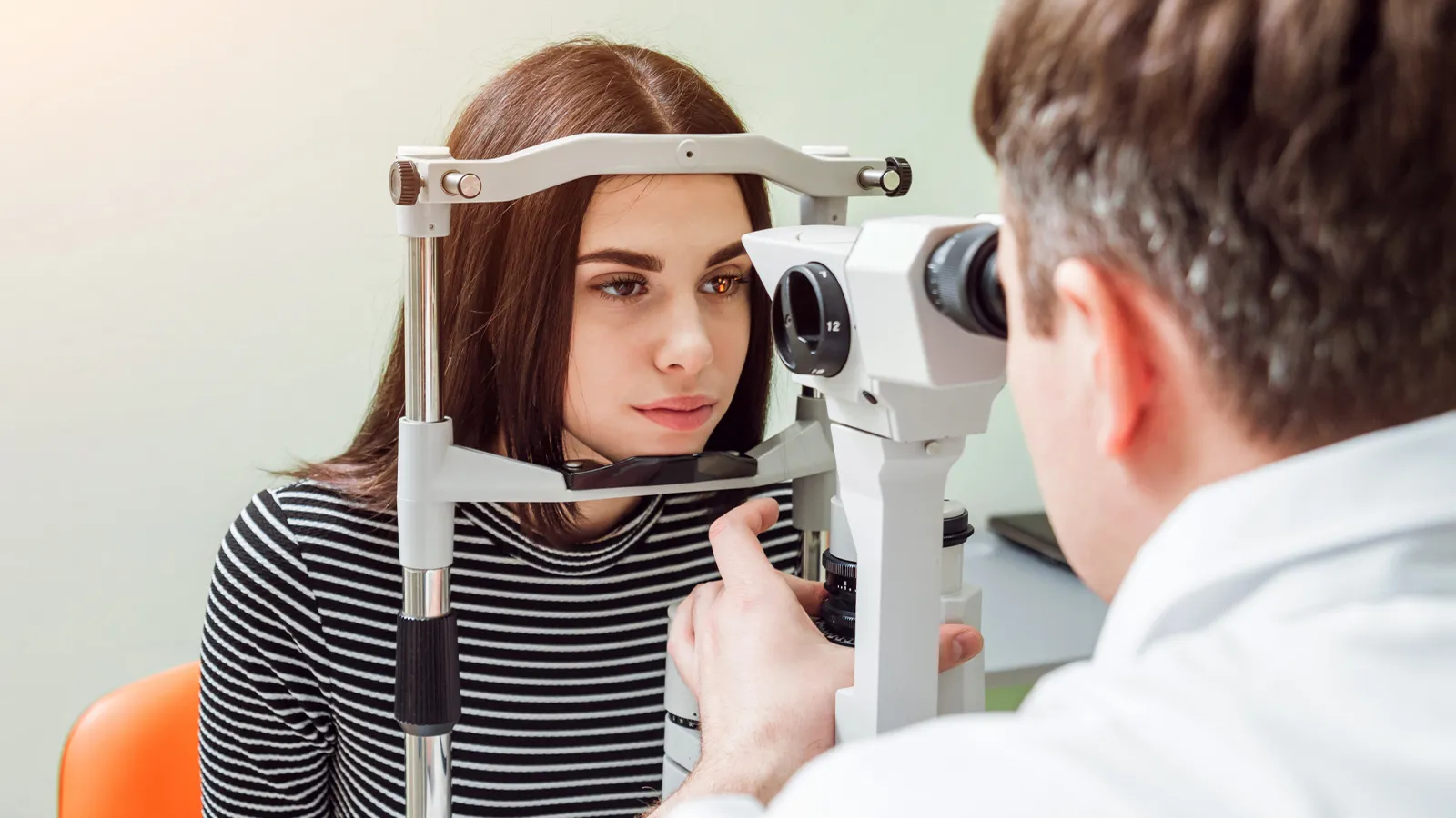 A woman with dark hair is undergoing a professional eye examination with an optometrist using a slit lamp microscope.