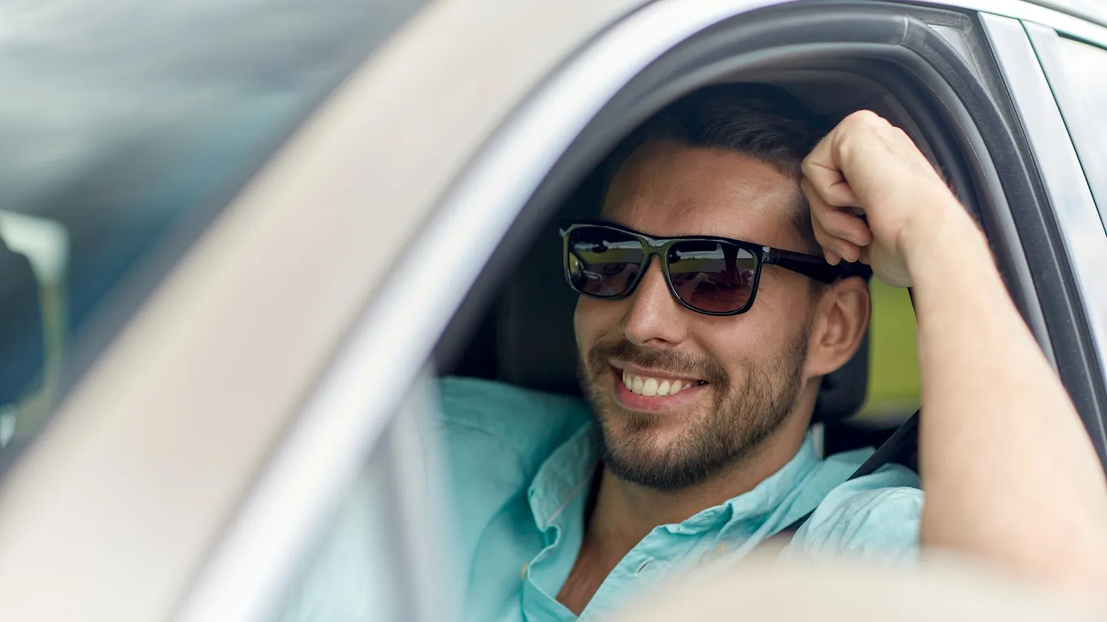 A man is sitting relaxed in his car, leaning his arm on the window frame with a cheerful smile. He wears black sunglasses for clear vision and more comfortable driving.