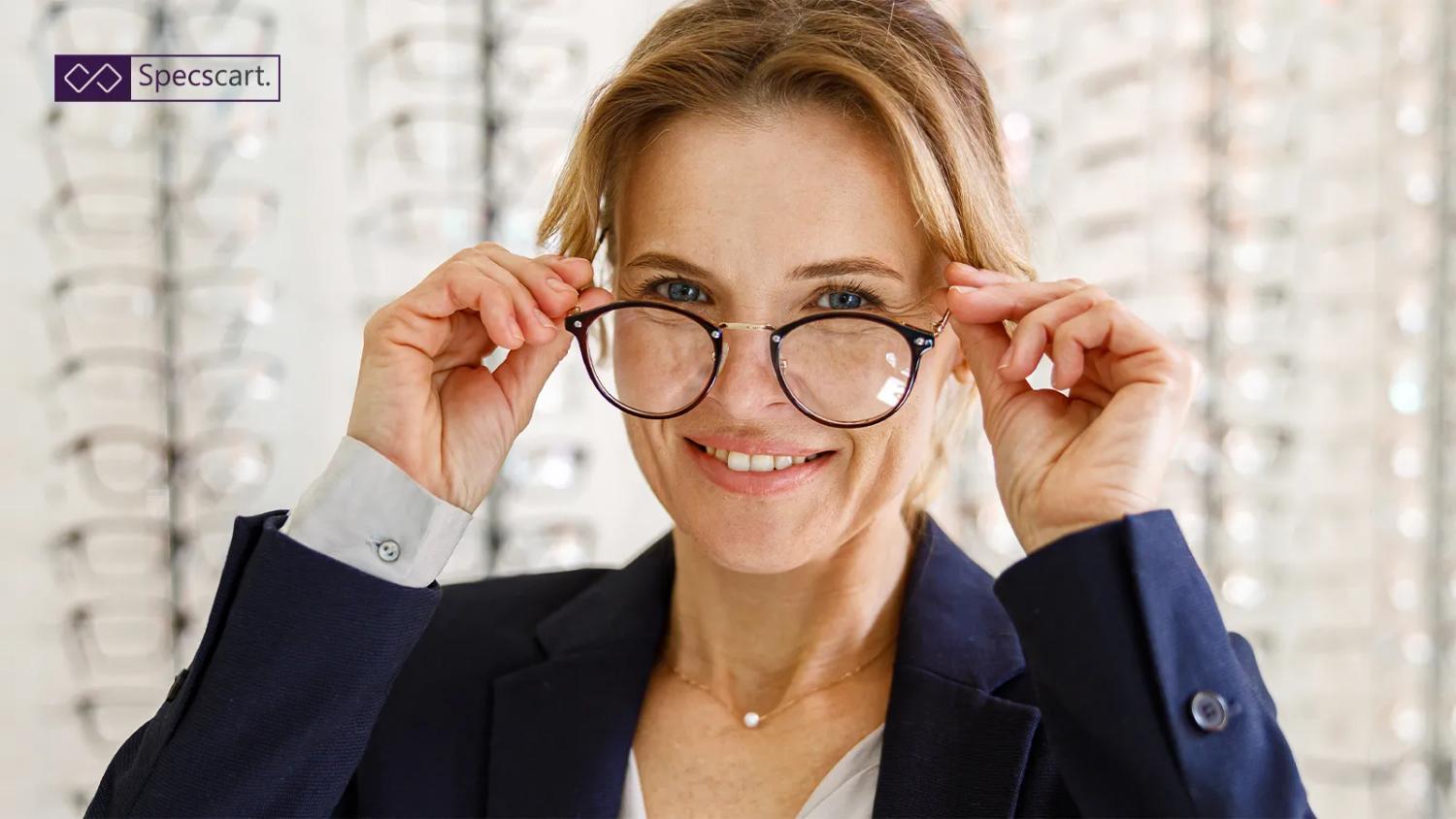 A smiling woman wearing a dark blazer tries on a pair of stylish round glasses with black and gold frames in front of a display rack in an optical store.