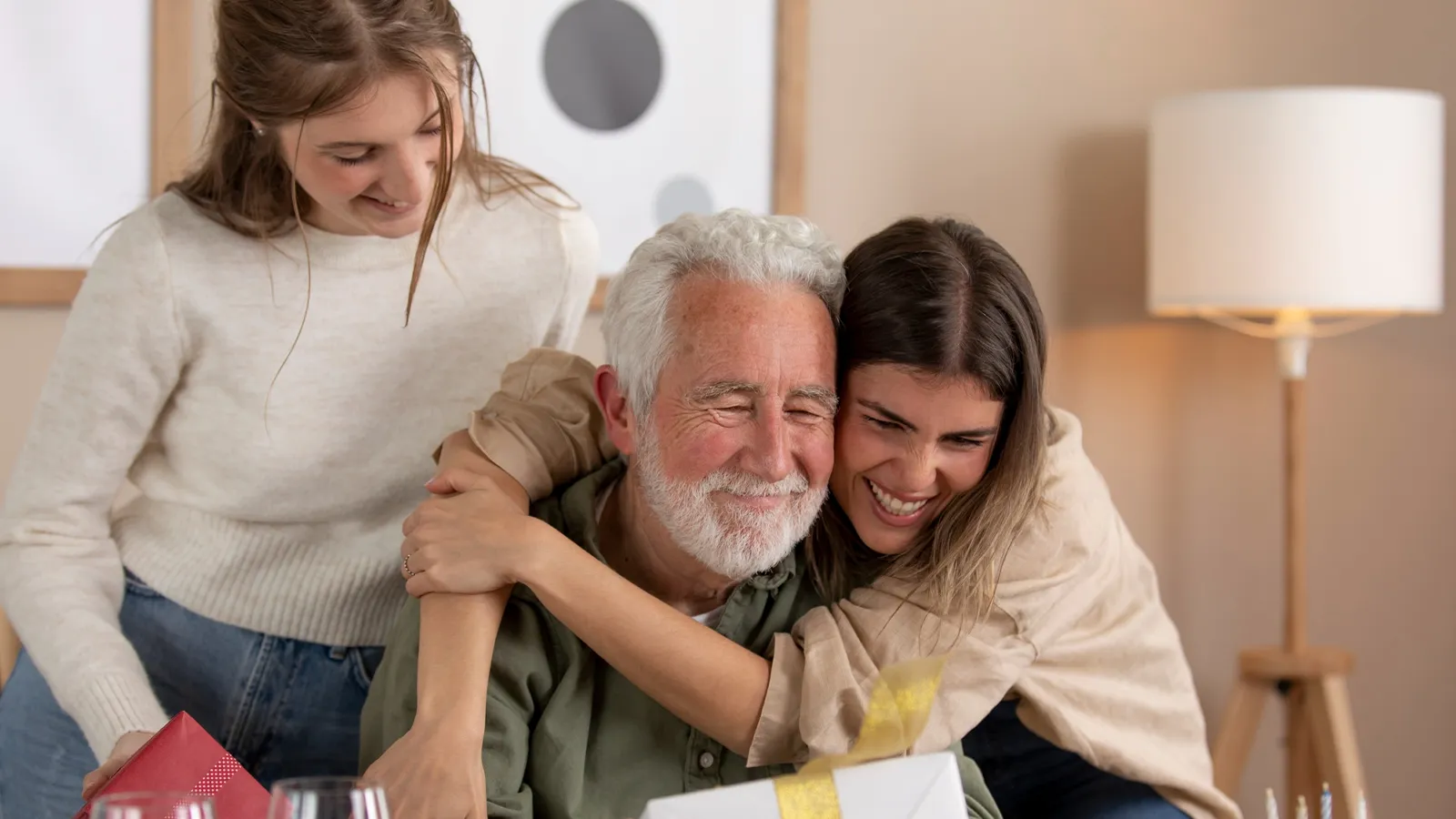A picture featuring an old man with two of his daughters spending time in the living room. One of them is hugging him and the other one stands right beside them. All three of them are wearing winter clothes and have a smiling expression.