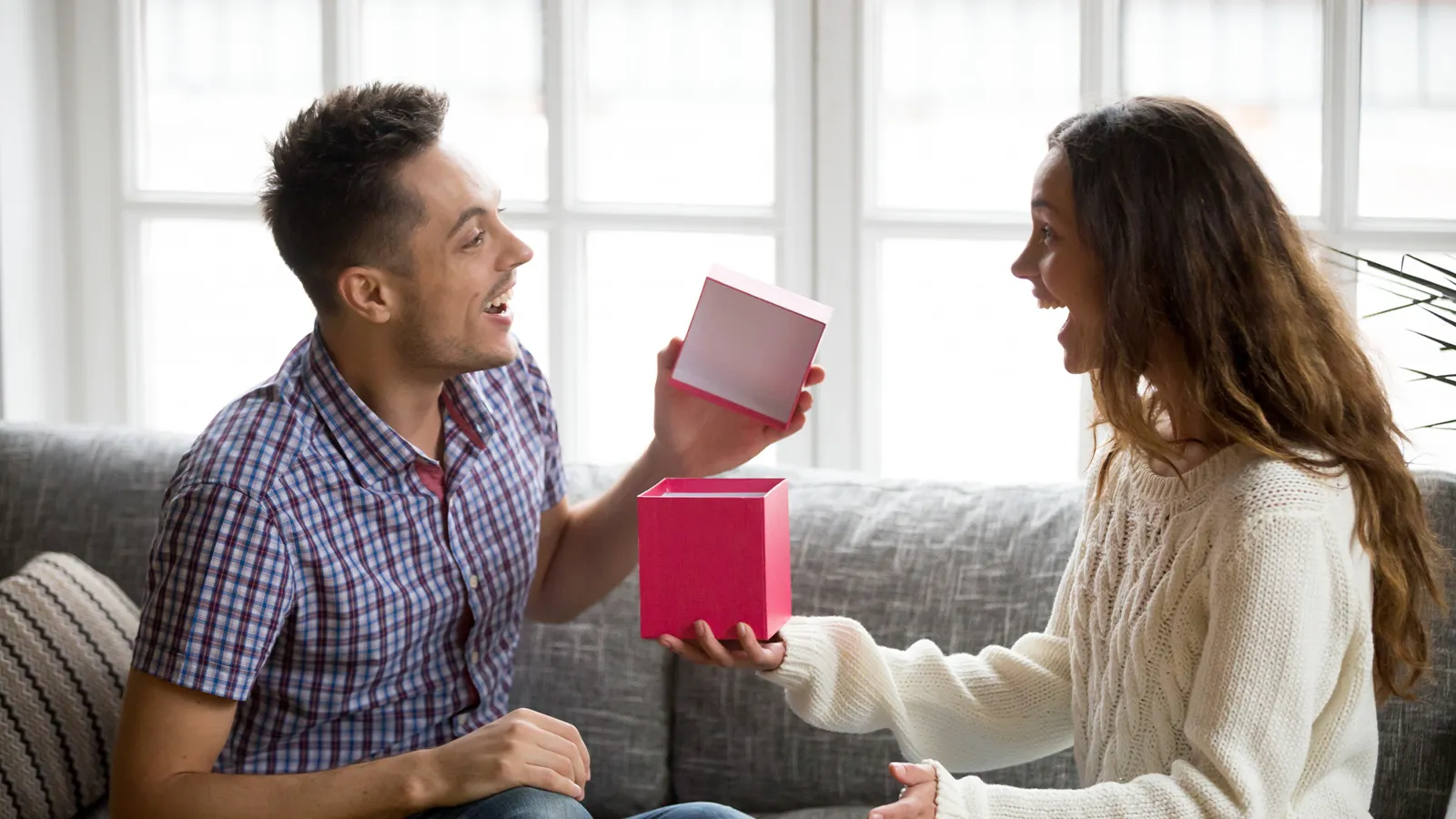 An image featuring a brother and sister giggling together, sitting on a sofa. Both of them are laughing and sharing gifts and laughing.