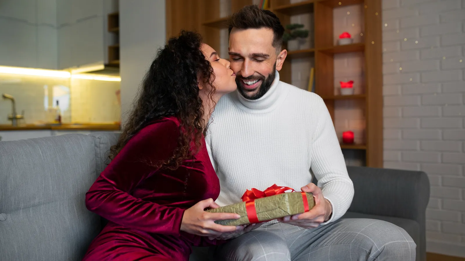 A picture featuring a woman wearing a maroon velvet dress kissing her husband and giving him a gift. Both of them are sitting on a sofa and look in love. They have a smiling expression.