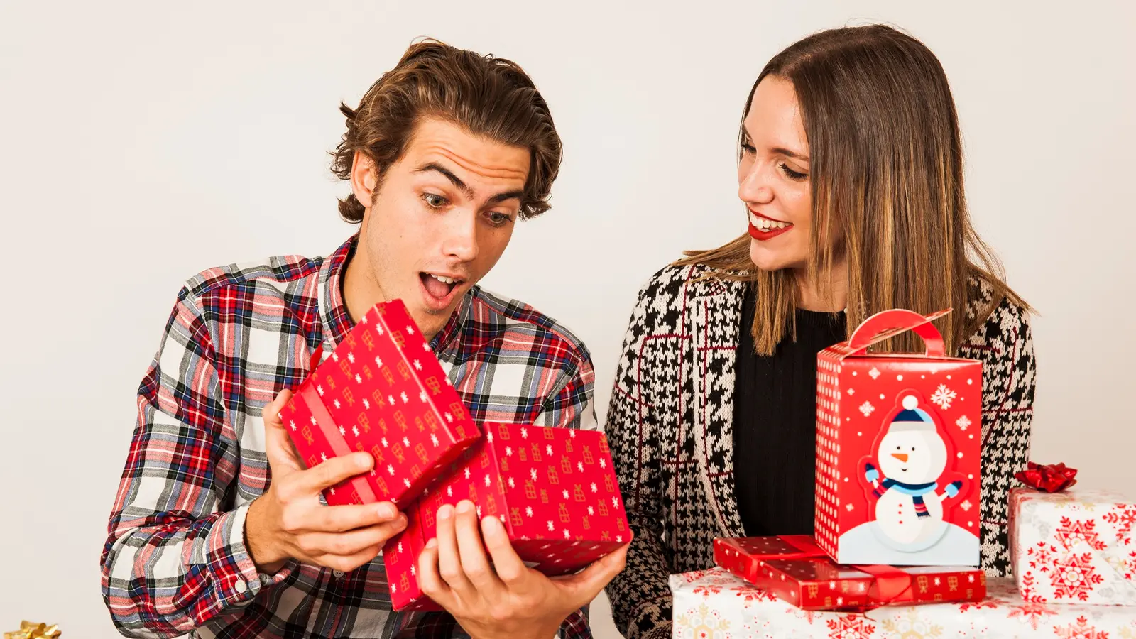 An image with a man and woman sharing gifts. The man has a shocked expression on his face, whereas woman is smiling and looking at his expression. The gifts are packed in red coloured gifting paper.
