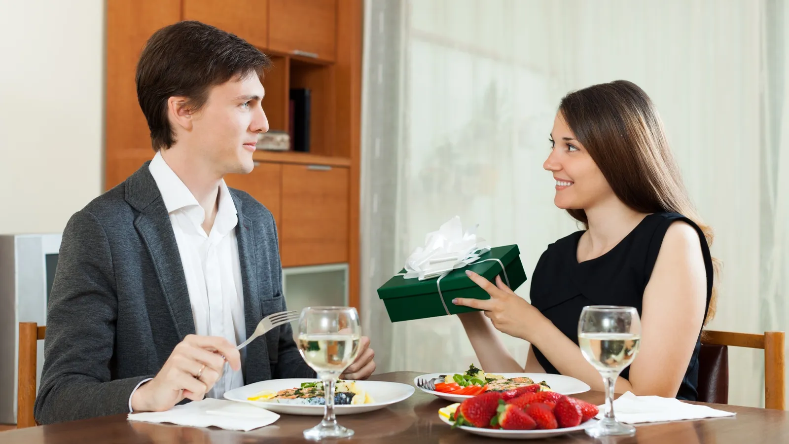 The image features a man and a woman sitting on a dining table and having food and fine. In the picture woman is wearing a black dress and her hair is open while she gives a gift to the male.