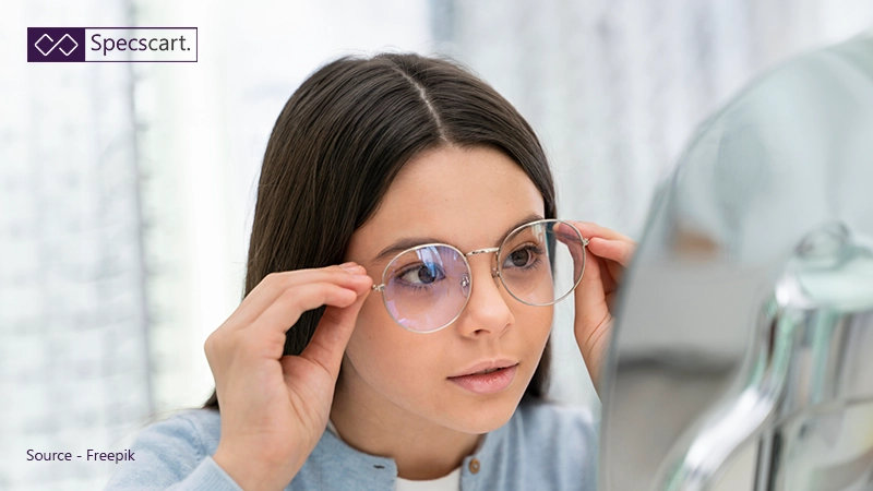 A young girl adjusting round metal eyewear in a mirror, focusing on the bridge and nose pads of the glasses.