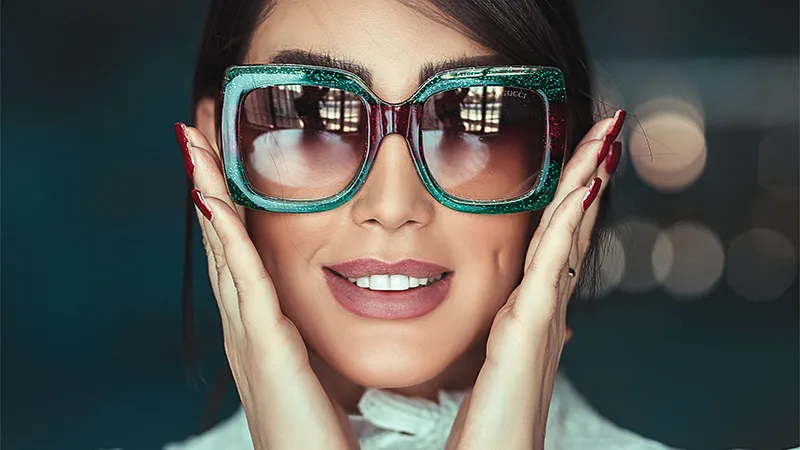 Woman wearing massive green glitter square sunglasses with red temple details and gradient lenses.