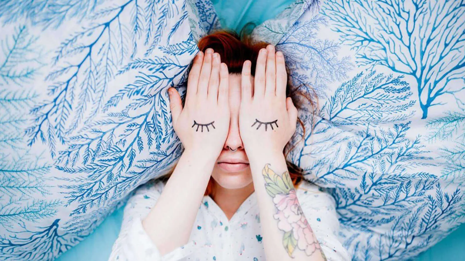 A person lying in bed with their hands covering their eyes, illustrating sleep disruption and insomnia caused by excessive exposure to blue light.