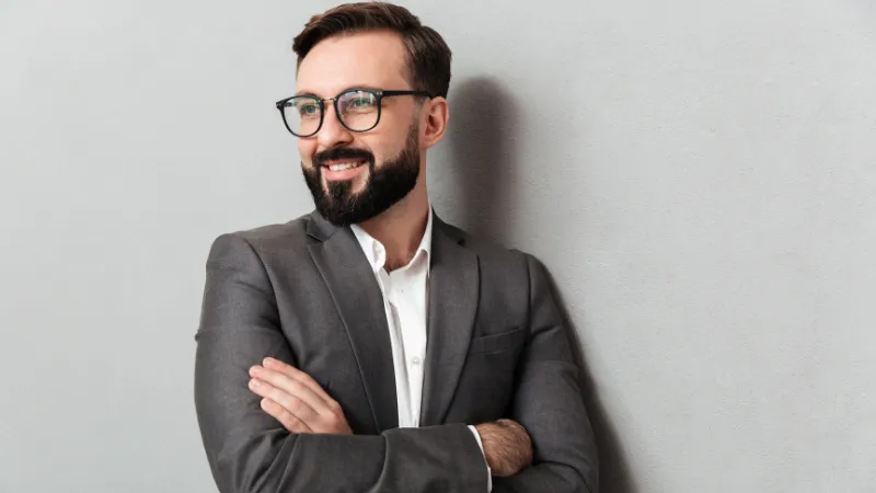 A man with a modern hairstyle, looks intently with a smile, wearing a light grey shirt and stylish, round black-rimmed glasses.