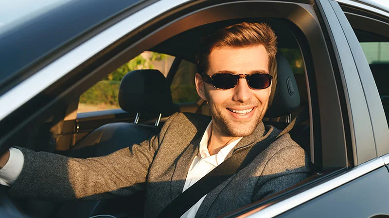A smiling man is driving a car, wearing a seatbelt and a grey tweed blazer. He looks to the side while wearing small, dark tinted Transitions XTRActive lenses in bright sunlight.