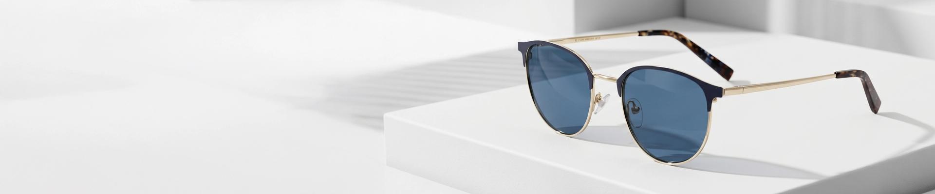 Brow Bar Sunglasses Close-up of sleek brow bar sunglasses with dark-tinted lenses resting on a white geometric block against a soft, minimalist white background.