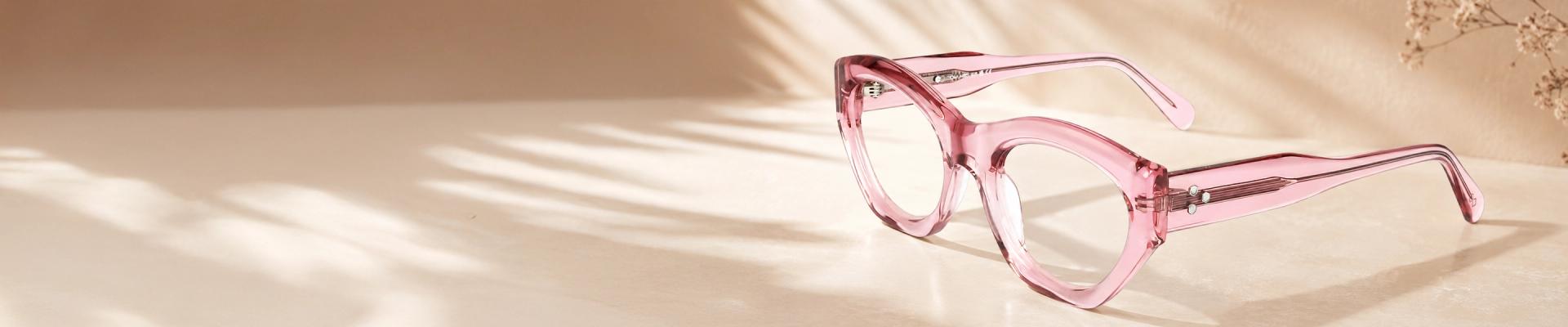 Pink Glasses Translucent crystal pink round glasses frames resting on a reflective soft pink background, showcasing a trendy and chic monochrome aesthetic.