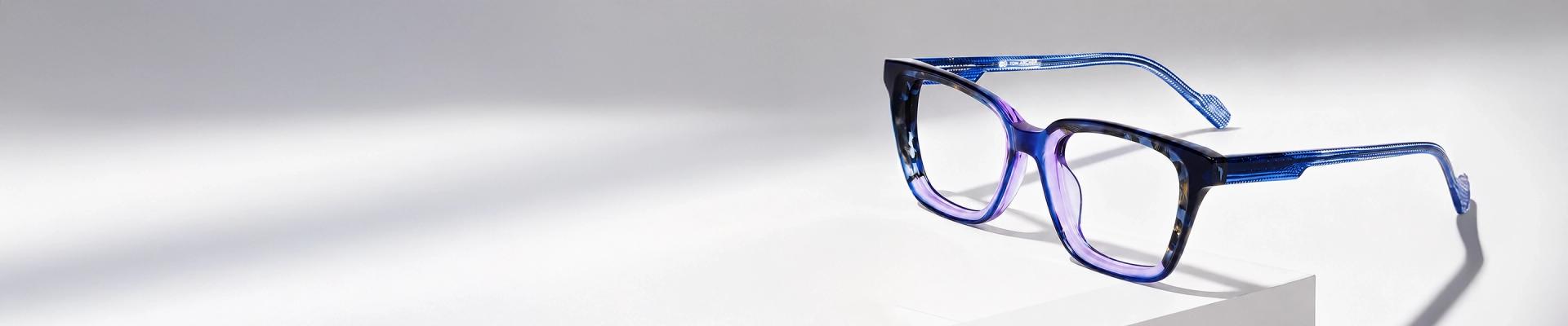 Purple Glasses Image showing a sleek pair of purple cat-eye prescription glasses with thin silver metal detailing on the sides, resting on a white cylindrical display stand, advertising purple glasses.