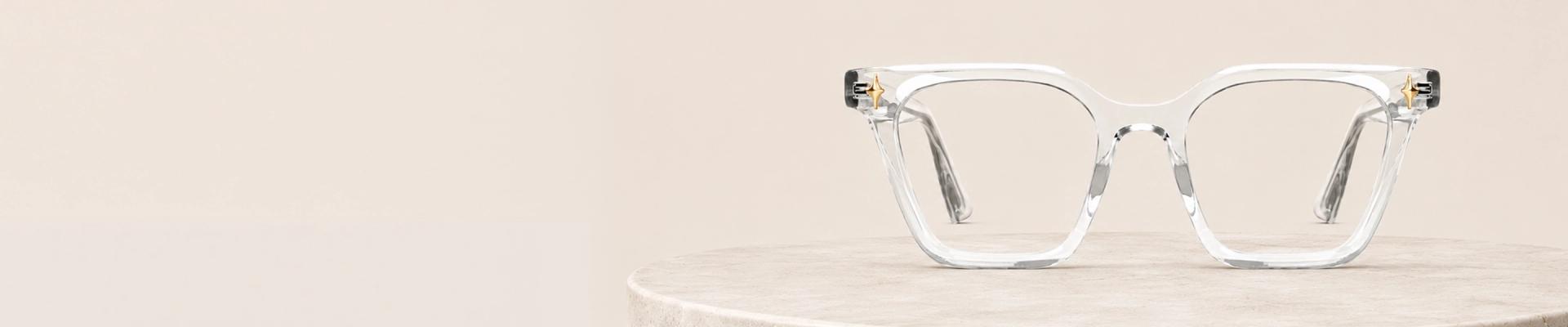 Clear Glasses The image features a close-up of clear acetate eyeglasses in a chunky rectangular frame that is placed on a white stand against a light grey background. The image has content that says Clear Glasses. It also has a discount offer by Specscart and Trustpilot reviews.