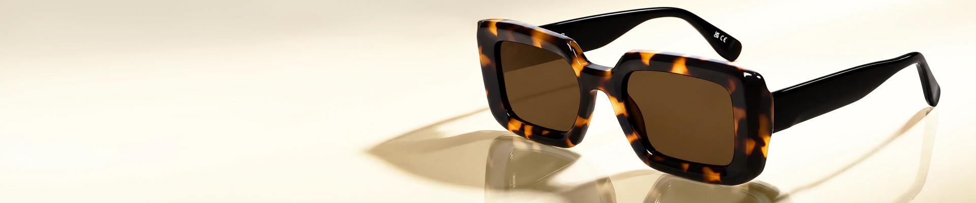 Tortoise Shell Sunglasses Smiling woman with hair tied back wearing chic rectangular tortoiseshell sunglasses with dark lenses, against a soft light blue gradient backdrop.