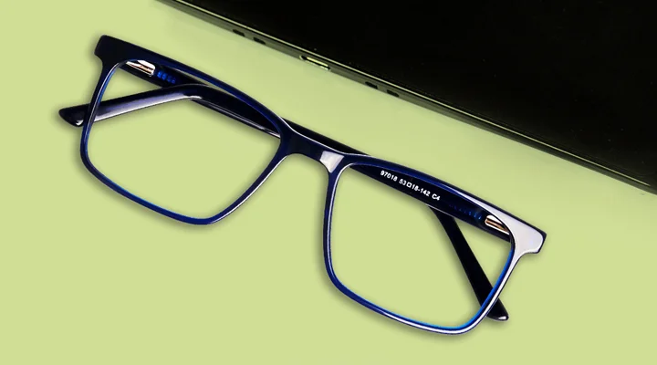 Blue rectangular glasses frames placed on a lime green desk next to a laptop, showcasing stylish and affordable eyewear for daily work.