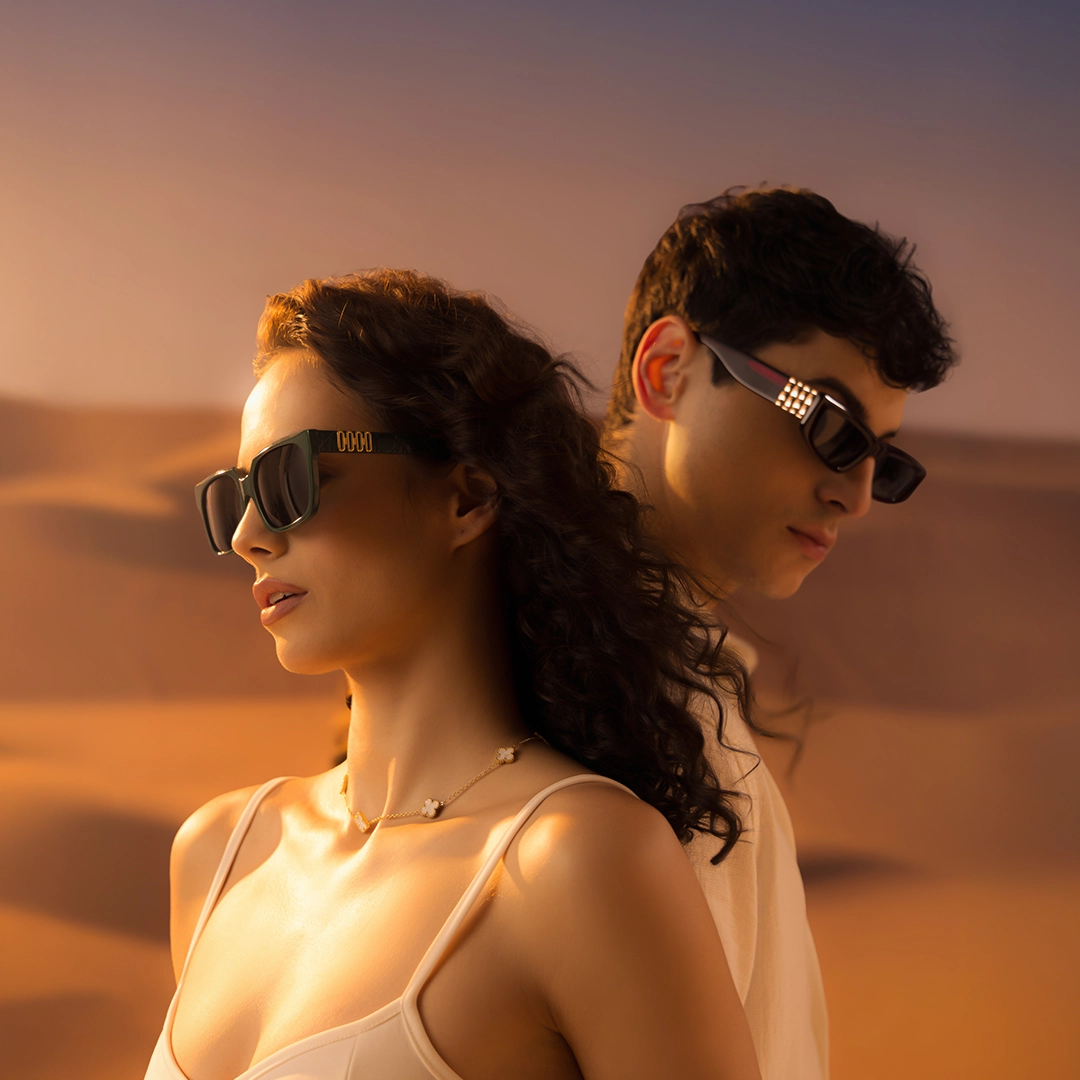 A man and woman pose back-to-back wearing premium designer sunglasses with gold temple details, set against a vast, golden desert dune landscape.