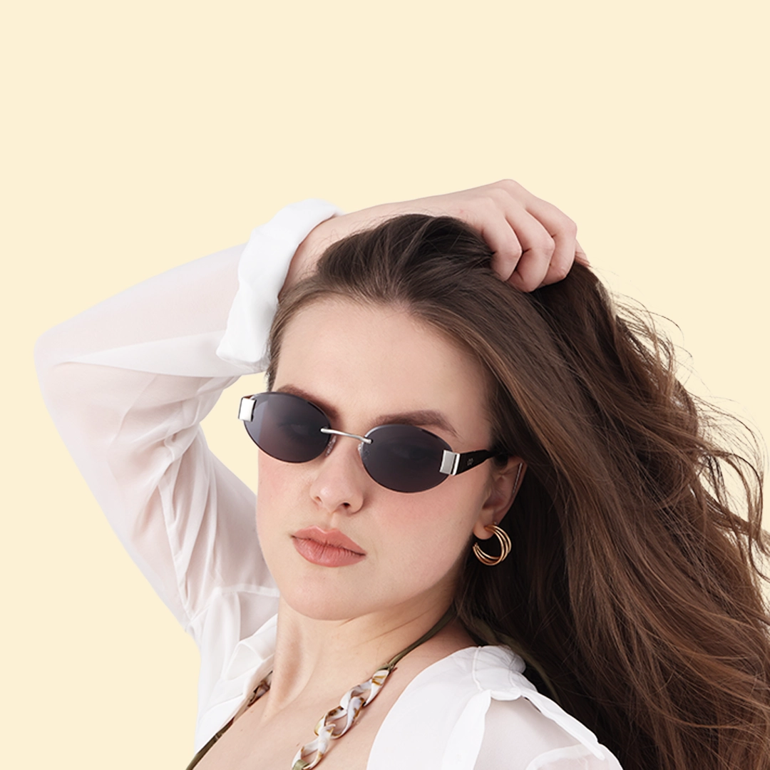Rimless Sunglasses A woman with wavy hair posing in small rimless oval sunglasses with dark lenses and gold hoop earrings against a minimalist light cream backdrop.