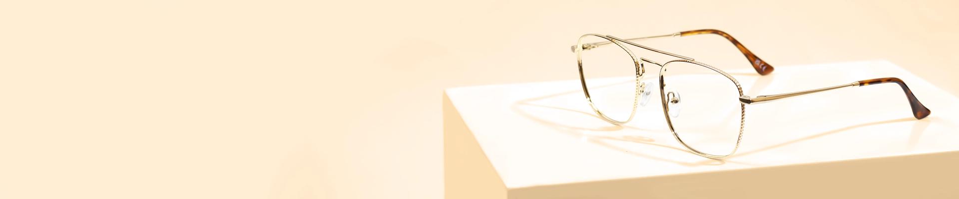 The banner features stylish glasses with gold-coloured metal rectangular frames, featuring intricate edges and tortoiseshell-patterned temple tips, presented on a white pedestal against a light beige background. The left side of the banner showcases promotional text for Gold Glasses and an Excellent Trustpilot rating.