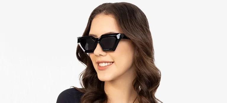 Geometric Sunglasses A woman with wavy brown hair wears thick black geometric sunglasses while smiling, set against a modern split background of light blue and white.