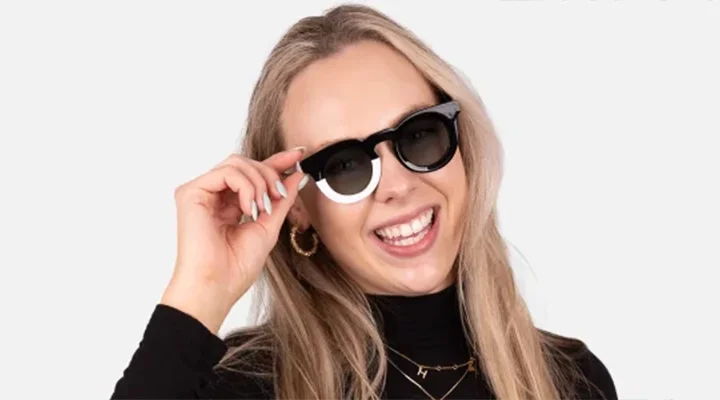 Funky Sunglasses A laughing blonde woman wearing unique two-toned black and white chunky round sunglasses, styled with a black turtleneck against a white backdrop.
