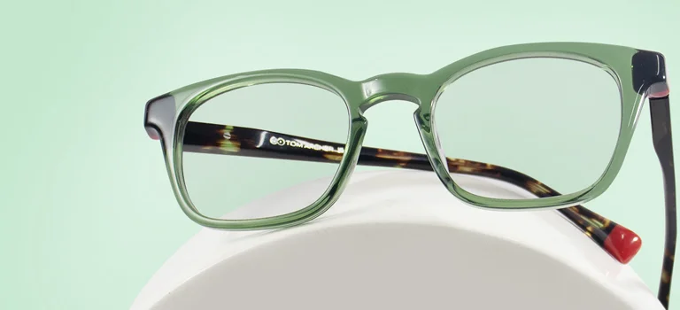 Green Glasses The image features a pair of green eyeglasses with black and tortoiseshell temples. It has a rectangular shape and is placed over a white stand against a light green background. It also has text saying Green Glasses, discount offer and Trustpilot Reviews.