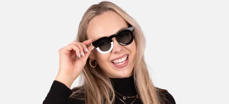 Funky Sunglasses A laughing blonde woman wearing unique two-toned black and white chunky round sunglasses, styled with a black turtleneck against a white backdrop.