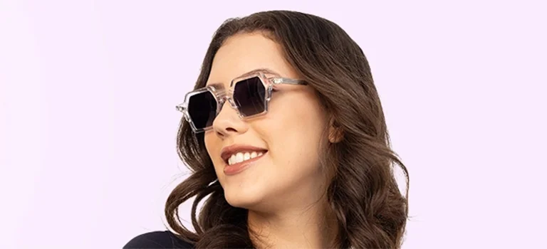 A smiling woman wearing modern hexagonal sunglasses with clear transparent frames and dark lenses, positioned against a soft lavender gradient banner.