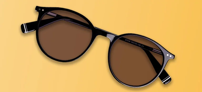 A pair of classic round sunglasses with black frames and brown tinted lenses, laid flat against a warm yellow and cream gradient background.