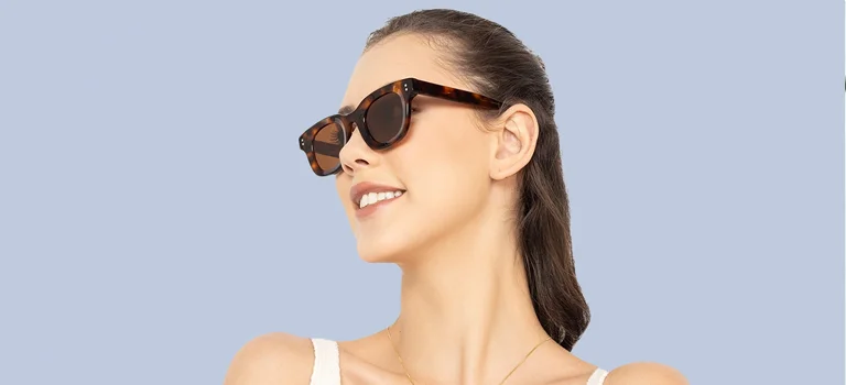 Smiling woman with hair tied back wearing chic rectangular tortoiseshell sunglasses with dark lenses, against a soft light blue gradient backdrop.