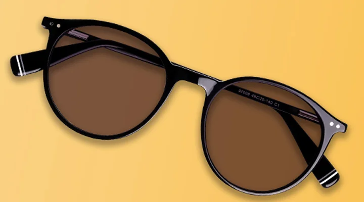 A pair of classic round sunglasses with black frames and brown tinted lenses, laid flat against a warm yellow and cream gradient background.