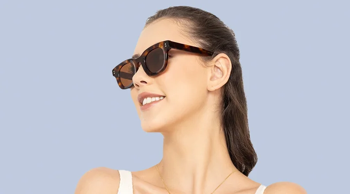 Smiling woman with hair tied back wearing chic rectangular tortoiseshell sunglasses with dark lenses, against a soft light blue gradient backdrop.