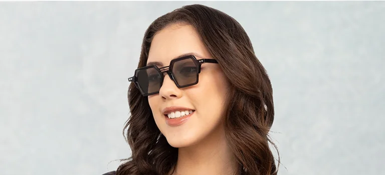 A smiling woman wearing black hexagonal sunglasses with a unique triple-bridge design, posed against a textured light blue and grey banner background.