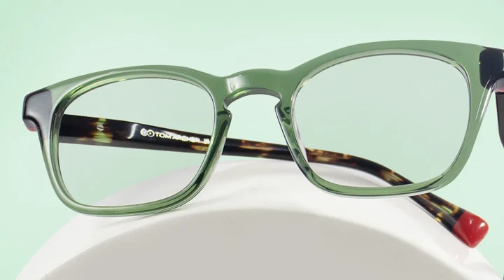 Green Glasses The image features a pair of green eyeglasses with black and tortoiseshell temples. It has a rectangular shape and is placed over a white stand against a light green background. It also has text saying Green Glasses, discount offer and Trustpilot Reviews.